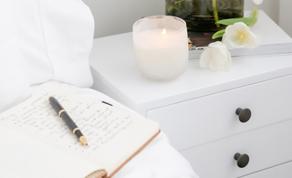 Open journal with handwritten notes and pen on a white bed beside a lit candle and fresh white flowers on a nightstand — a calming space for mindful journaling.