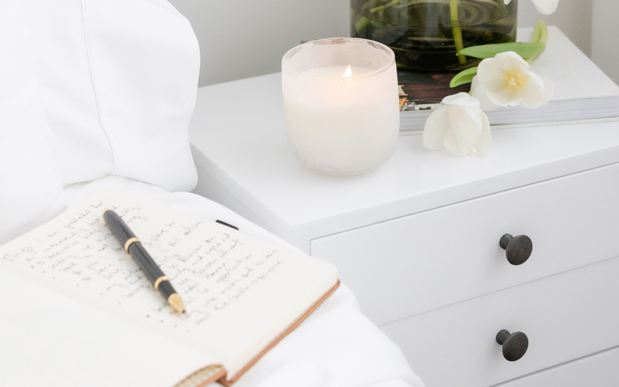 Open journal with handwritten notes and pen on a white bed beside a lit candle and fresh white flowers on a nightstand — a calming space for mindful journaling.
