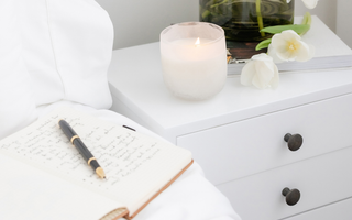 Open journal with handwritten notes and pen on a white bed beside a lit candle and fresh white flowers on a nightstand — a calming space for mindful journaling.