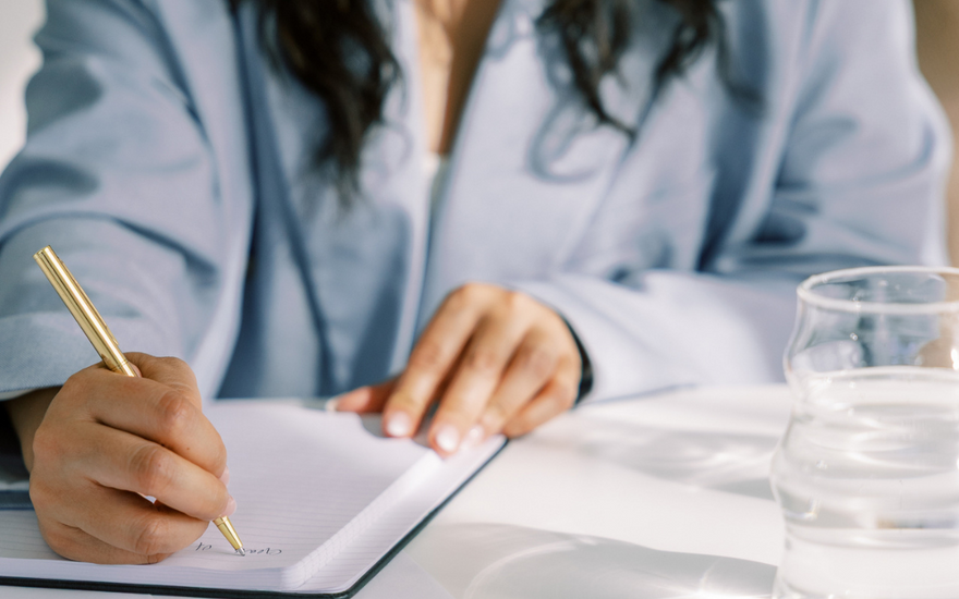 Woman writing in a journal with a gold pen at a bright desk, practicing reflective journaling for a digital detox.