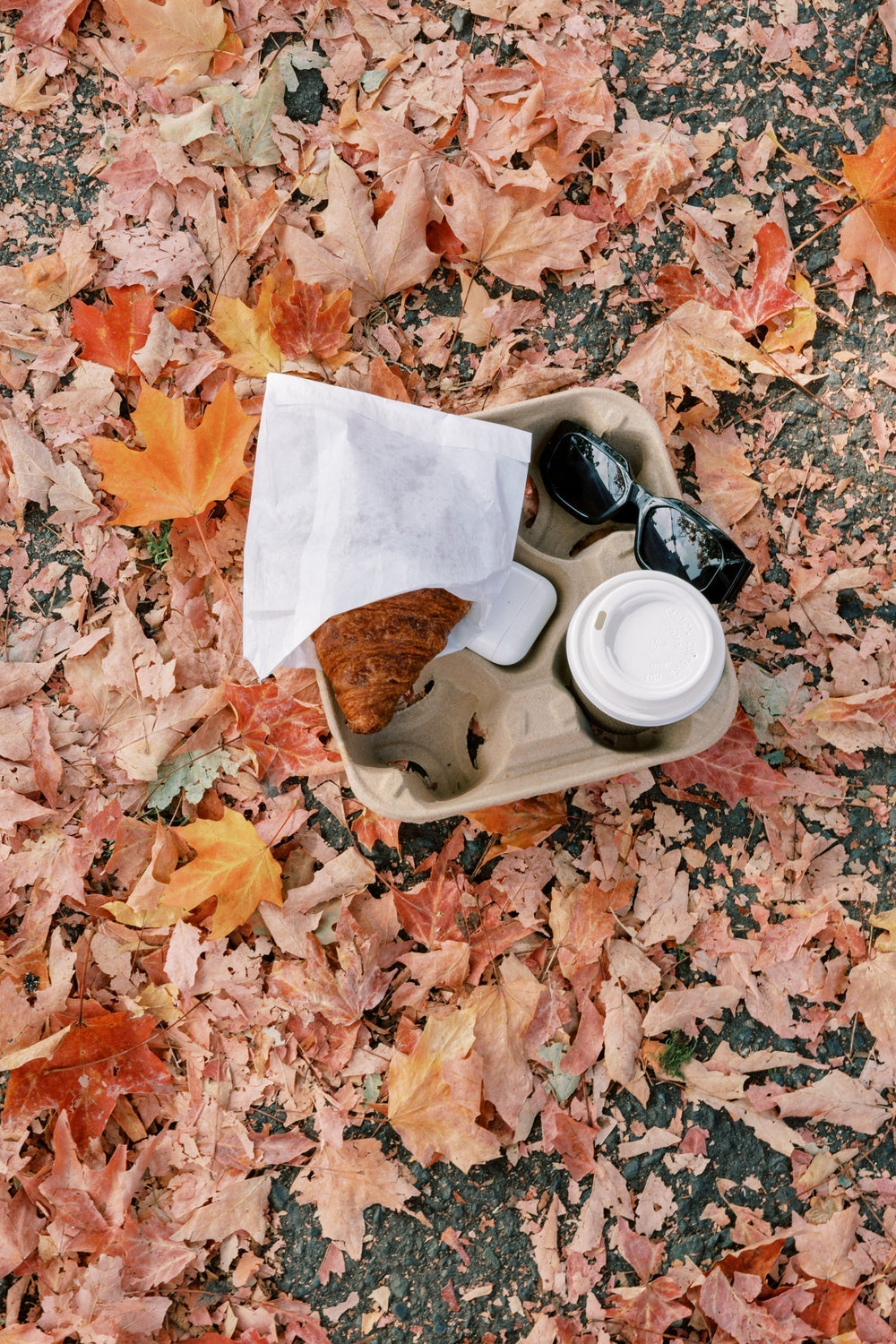 A takeaway tray with a croissant, coffee cup, sunglasses, and earbuds rests on a blanket of fallen autumn leaves, capturing a cozy fall moment on the go.
