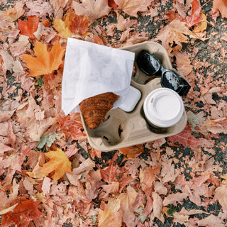 A takeaway tray with a croissant, coffee cup, sunglasses, and earbuds rests on a blanket of fallen autumn leaves, capturing a cozy fall moment on the go.