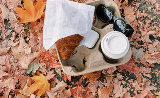 A takeaway tray with a croissant, coffee cup, sunglasses, and earbuds rests on a blanket of fallen autumn leaves, capturing a cozy fall moment on the go.