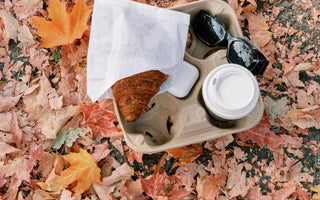 A takeaway tray with a croissant, coffee cup, sunglasses, and earbuds rests on a blanket of fallen autumn leaves, capturing a cozy fall moment on the go.