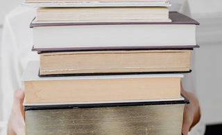 Smiling woman in white holding a tall stack of vintage hardcover books — symbolizing cozy, analog living and the joy of visiting a local library.