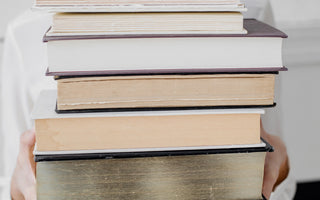 Smiling woman in white holding a tall stack of vintage hardcover books — symbolizing cozy, analog living and the joy of visiting a local library.