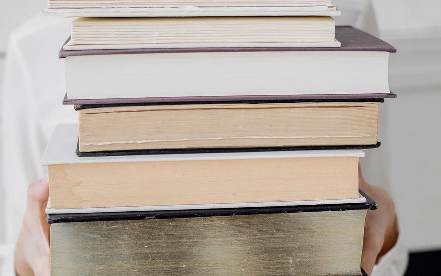 Smiling woman in white holding a tall stack of vintage hardcover books — symbolizing cozy, analog living and the joy of visiting a local library.